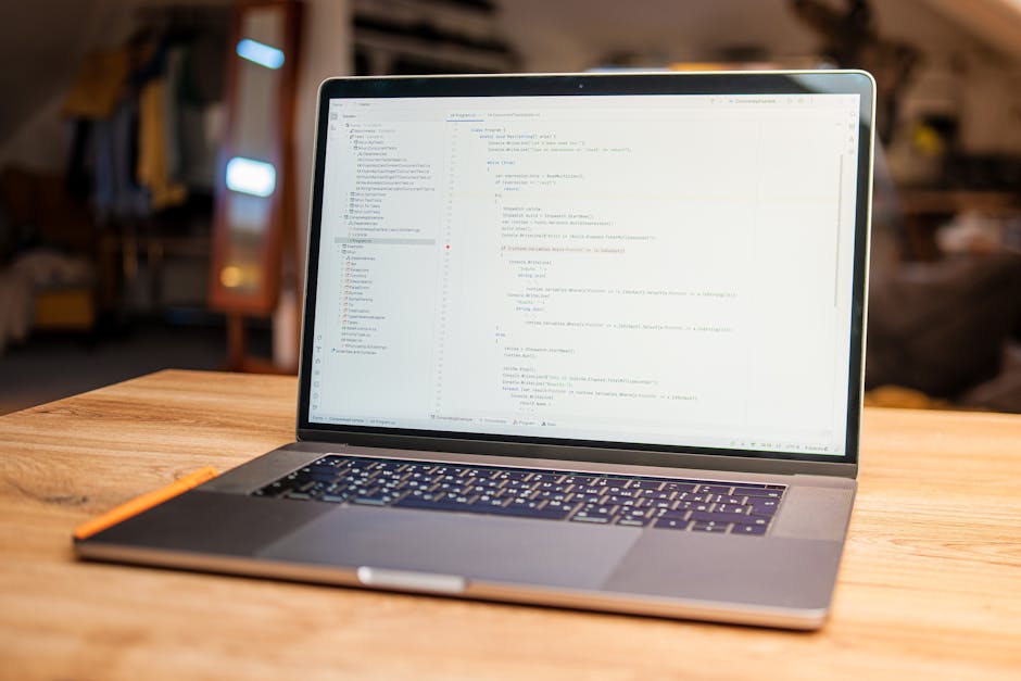 A close-up view of a laptop resting on a wooden table in an indoor setting, displaying lines of code on its screen with a light background and dark text. The laptop has a metallic finish and a black k