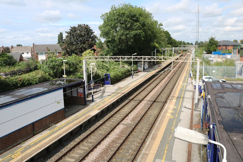 A view of Hackbridge Station platform from an elevated angle, showing the railway tracks running parallel to the platform. The platform features a small shelter with a dark, glossy roof, surrounded by