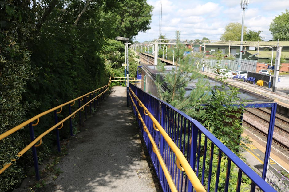 A view of Hackbridge Station platform from an elevated angle, showing the railway tracks running parallel to the platform. The platform features a small shelter with a dark, glossy roof, surrounded by